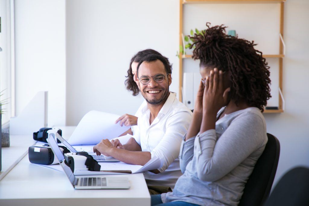 Joyful colleagues laughing during work. Happy office workers sitting at table with VR headsets and laptops. Technology concept